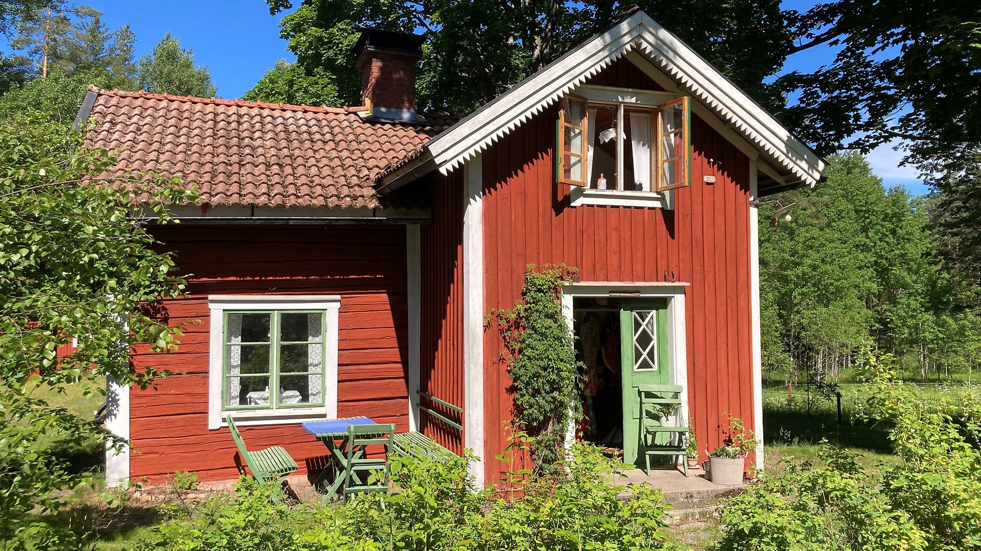 Ett rött trähus med vita knutar och en grön dubbeldörr som står halvt öppen till höger av huset. Framför ett fönster till vänster av huset står en grön utemöbel i trä. Framför huset finns grönskande buskar. 