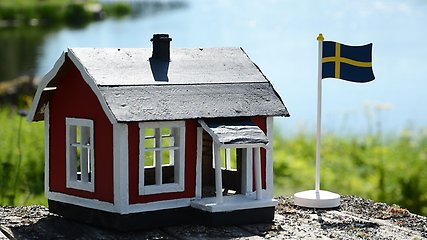 Ett miniatyrhus i rött trä och vita knutar. Huset står på en sten framför vatten och bredvid huset står en Sverige-flagga.
