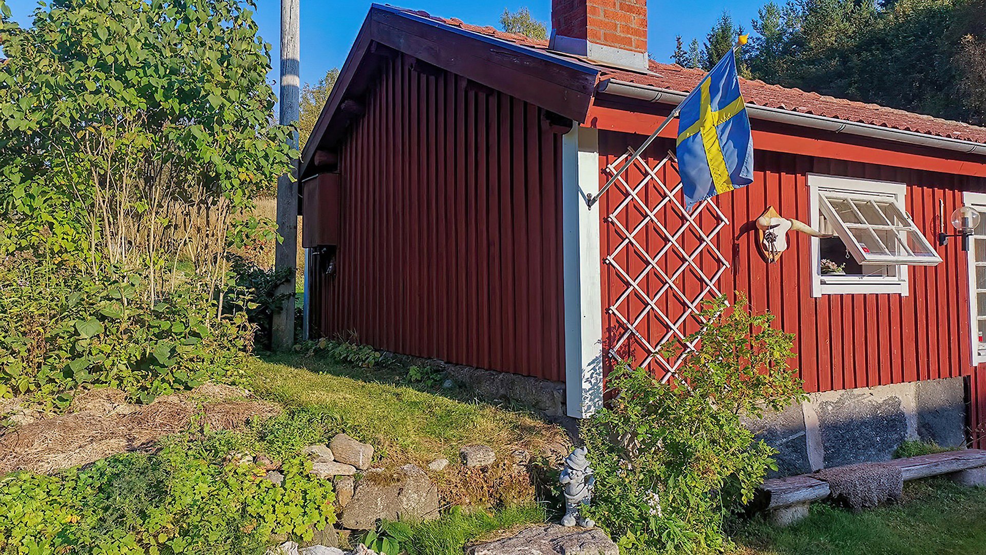 Ett rött trähus med vita knutar, ett öppet fönster och en Sverige-flagga. Till vänster om huset finns grönskande buskar.
