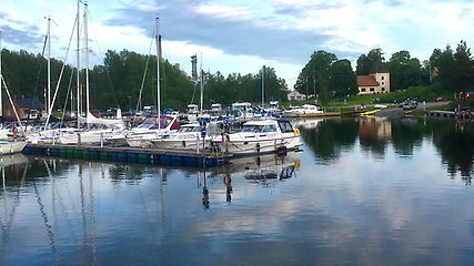 Hamn med många parkerade segel- och motorbåtar längs en brygga. Bakom båtarna syns flera byggnader, bland annat en kyrka till höger i bild.