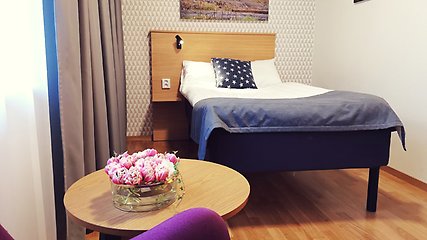 Hotellrum med en bäddad enkelsäng i centrum.  I förgrunden finns ett litet träbord med rosa tulpaner i en vas. Till vänster finns ett fönster med fördragna gardiner. 