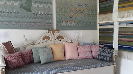Träsoffa med randig sittdyna på. På soffan finns ett flertal mönstrade kuddar i olika färger. På väggen bakom soffan hänger mönstrade tyger.