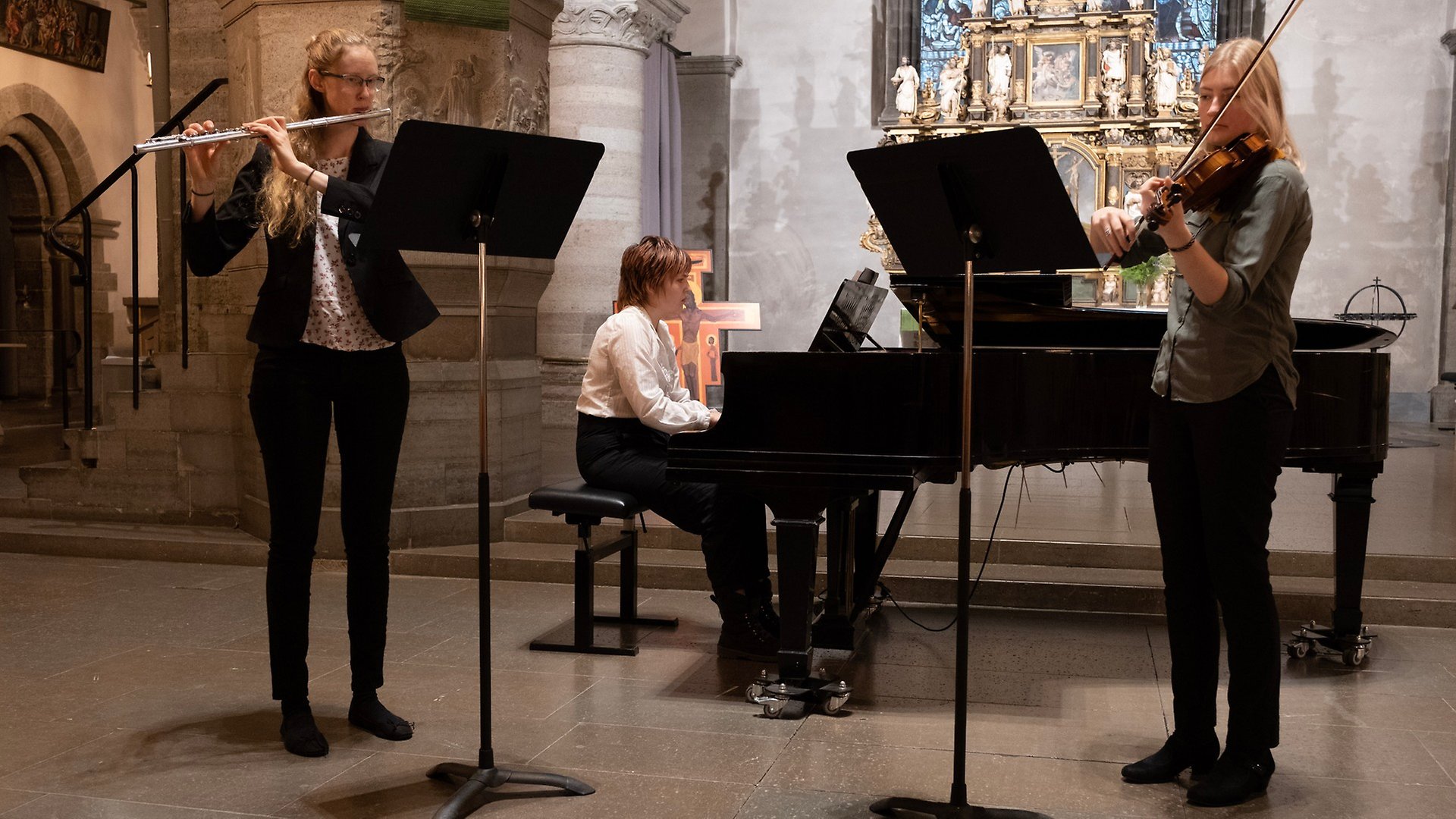 Tre tjejer spelar musik i en kyrka. Till vänster står en som spelar tvärflöjt och till vänster står en som spelar fiol. I mitten är det en som spelar piano vid en svart flygel.