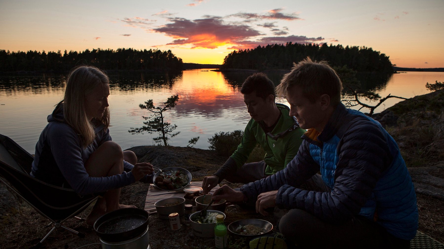 Ett sällskap på tre personer sitter vid lägereld på en ö. Solnedgång i bakgrunden.