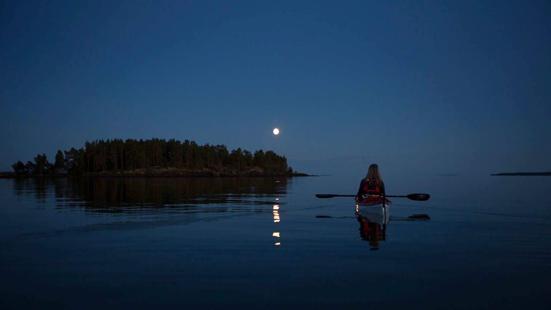 Liten måne speglar i i stilla vatten över en ö. En ensam paddlare i bilden.