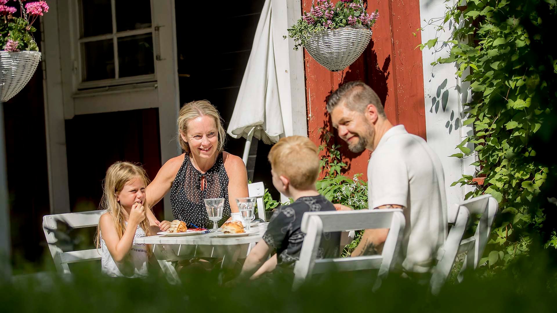 En kvinna, en man och två barn sitter runt ett cafébord i grönska och solsken och fikar. Alla tittar glatt på pojken. 