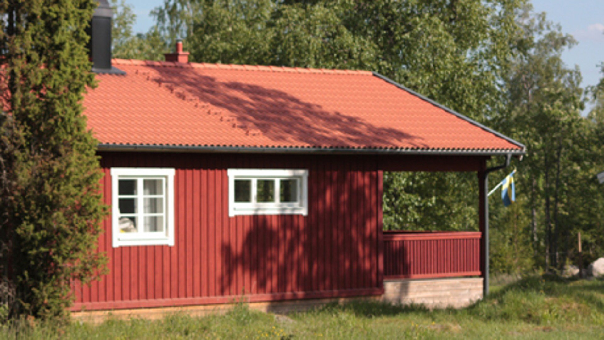 Sidovy av en röd stuga i trä. Till vänster står en enbuske. Stugan har två fönster på långsidan och en veranda med tak på ena kortsidan. Vid verandan händer en svensk flagga. Bakom stugan finns en skog. 