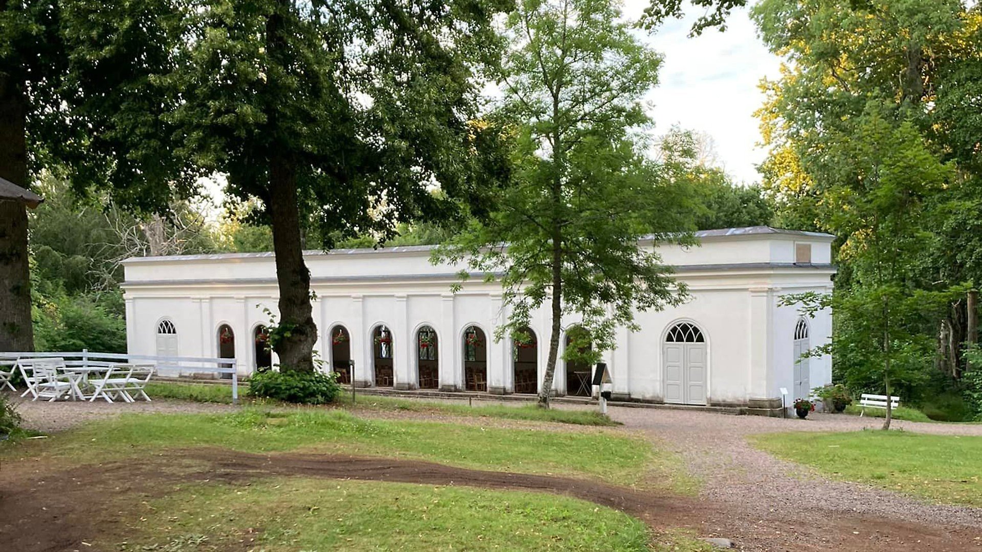 Högbrunnen vid Medevi Brunn, en avlång vit byggnad med höga valvfönster. I förgrunden finns flera grönskande träd och en gräsmatta. 