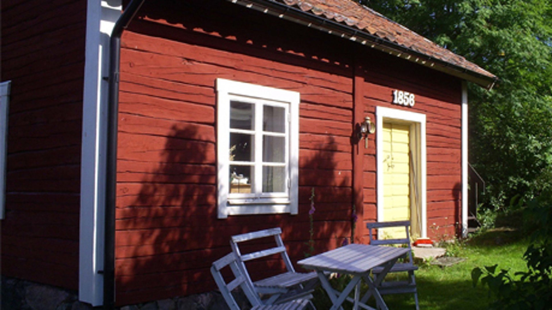 Ett rött trähus med vita knutar, ett fönster och en gul dörr. Ovanför dörren står det 1858 i vitt. Framför fönstret står vita utemöbler i trä. Solen lyser på huset och skuggan av ett träd syns. 