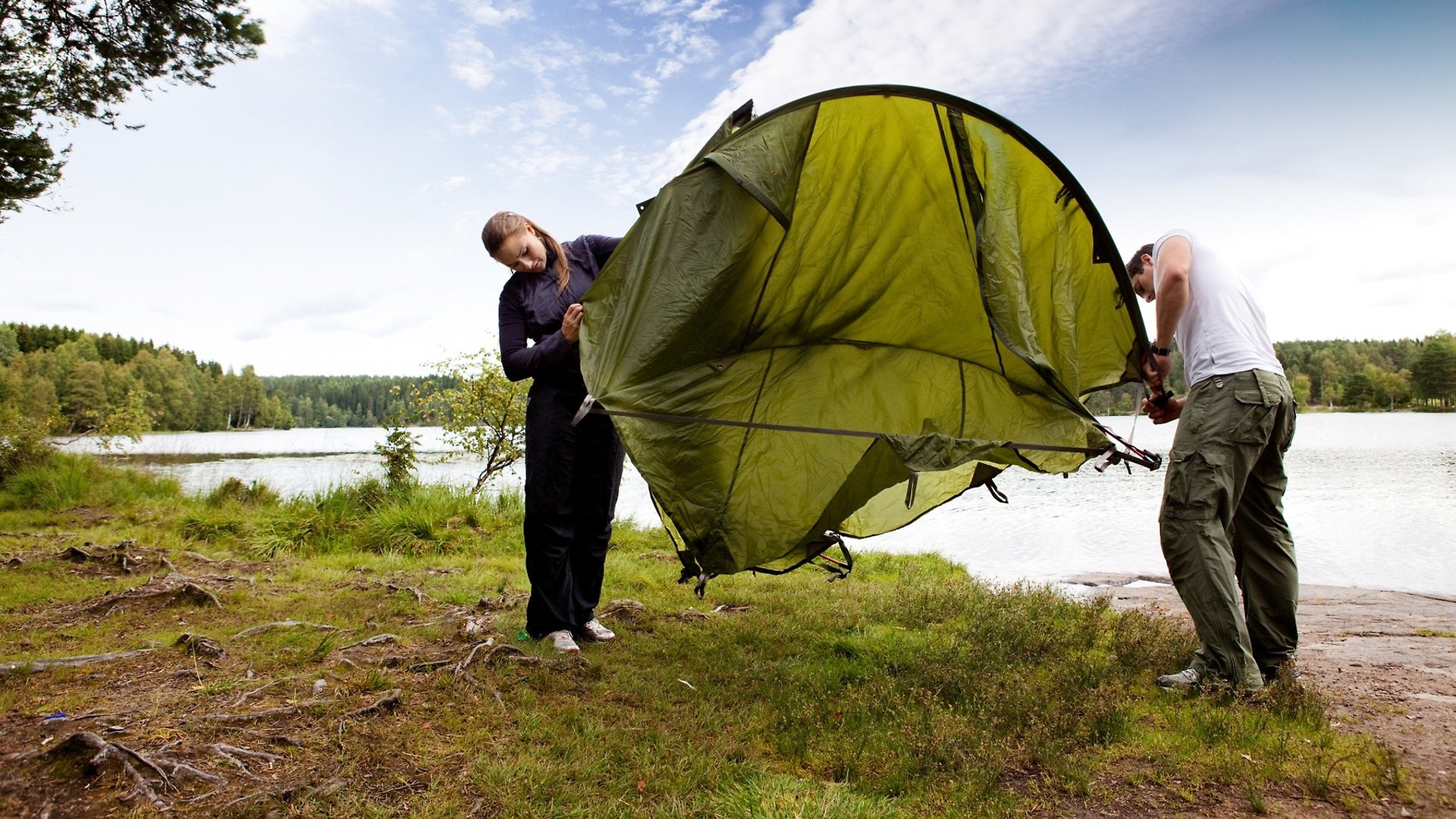 Två personer som sätter upp ett tält vid en sjö ute i skogen.