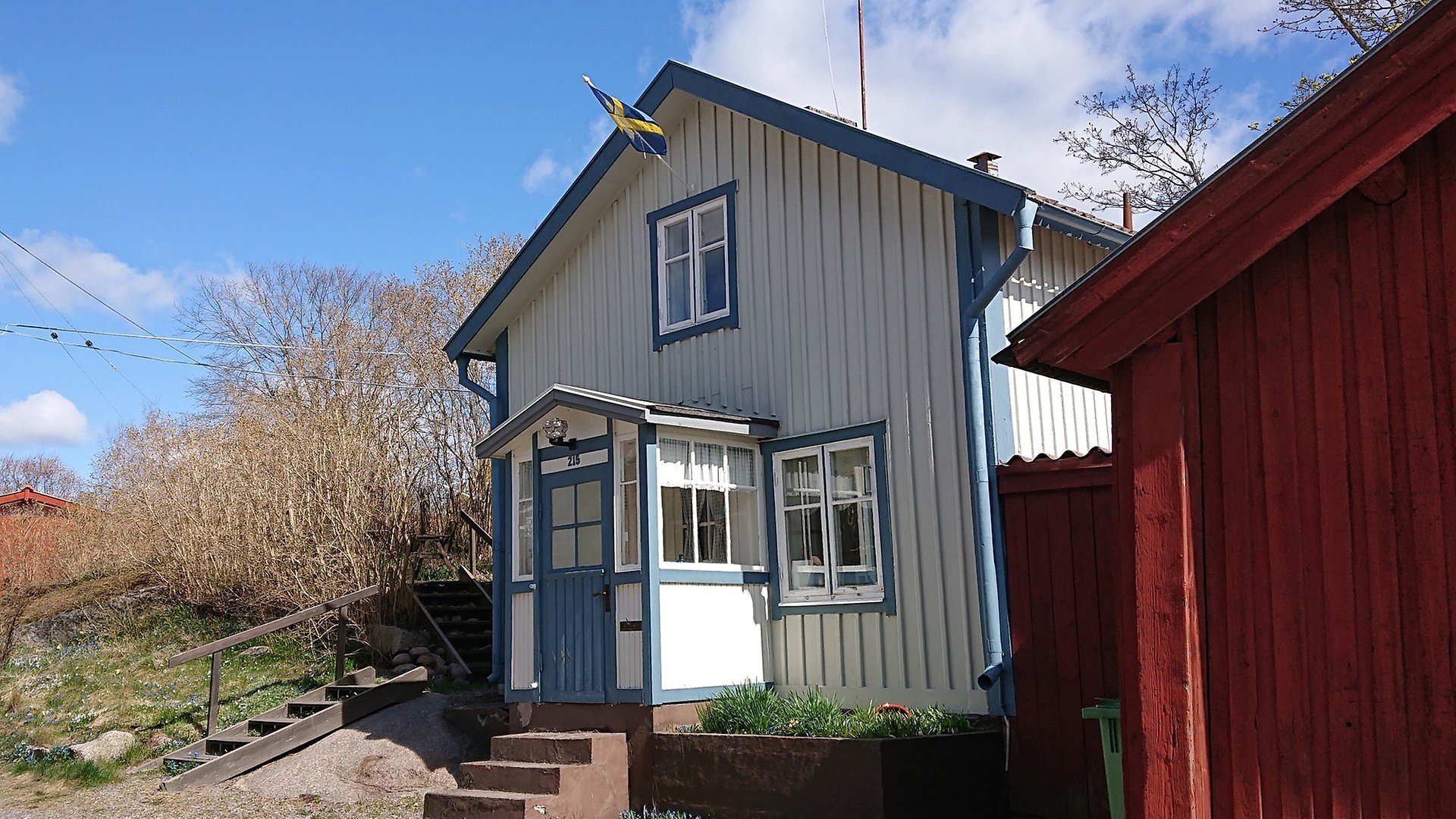 En bild tagen på kortsidan av ett vitt trähus med blåa knutar. Över ett fönster hänger en Sverige-flagga. Bakom huset syns en blå himmel och träd utan löv. 