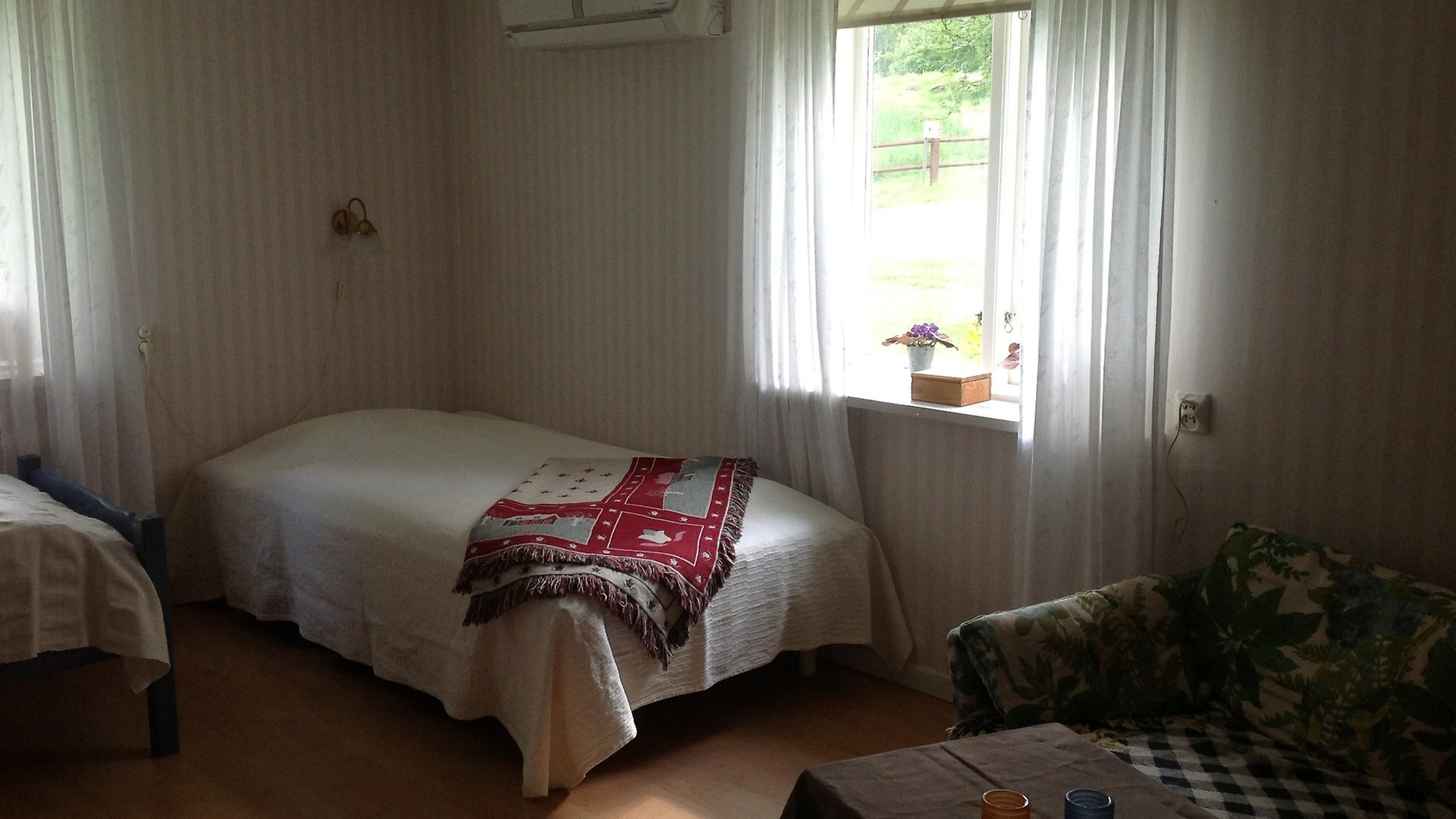En säng i ett hörn av ett rum med ett fönster på båda väggarna. Sängen är bäddad med vitt överkast och en röd filt. I utkanten av bilden finns hörnet av en soffa. 