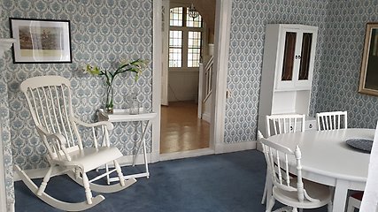 Ett rum med stormönstrad tapet i blått och vitt. I högra hörnet syns ett runt matbord och tre stolar i vitt. I vänstra hörnet syns en vit gungstol och ett sidobord med vita liljor i en vas. Mitt på väggen är en dörröppning ut mot ett stort fönster i rummet intill.