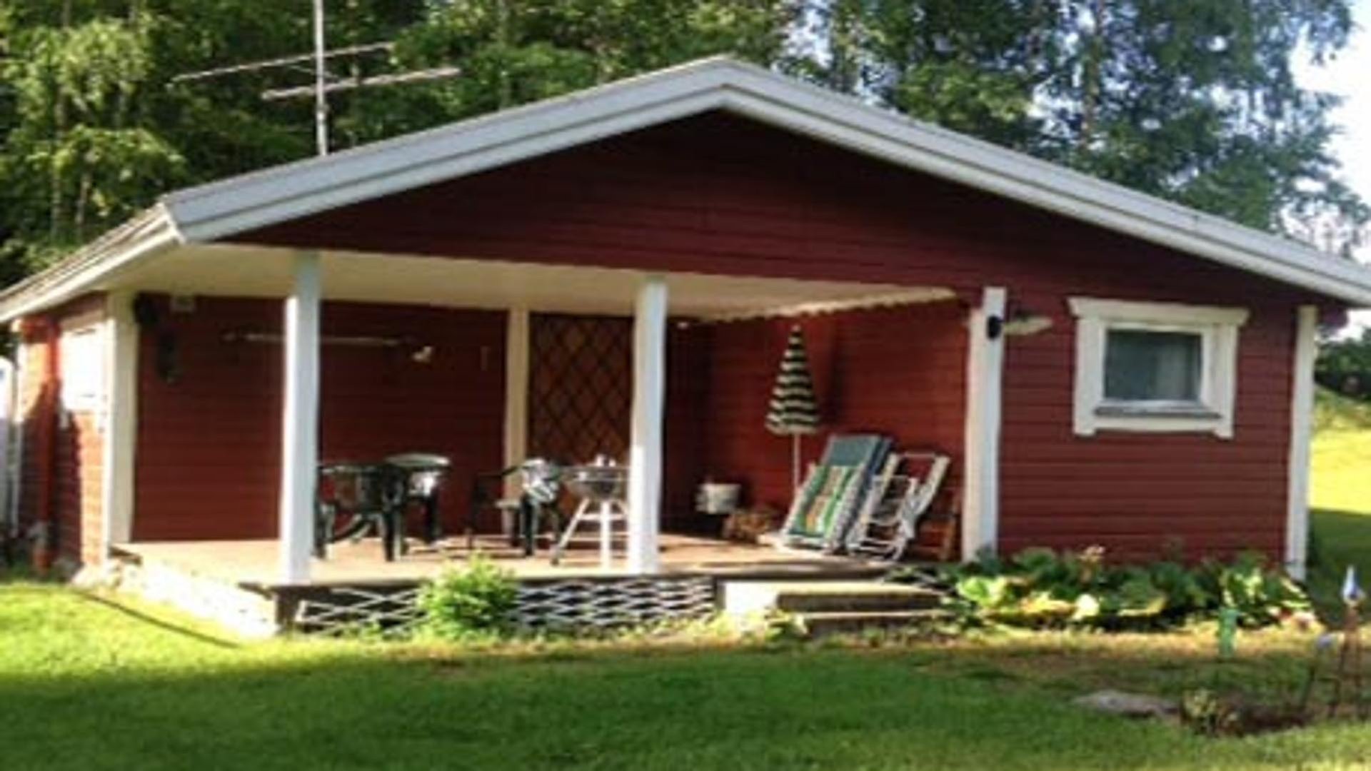 Ett rött trähus med vita knutar. Till höger finns en altan med grill, utemöbler och parasoll. 