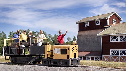 En man i röd tröja kör ett litet gult tåg på en räls. Han pekar mot något och fyra personer som står på i den öppna vagnen tittar åt hållet föraren pekar. Alla ler. I bakgrunden syns ett rött trähus med många fönster.
