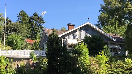 Ett grått trähus bakom grönskande buskar. Till vänster om huset syns ett vitt staket och en flaggstång utan flagga.