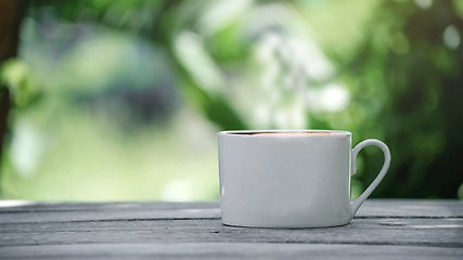 En vit mugg med kaffe på ett träbord och grönska bakom. 