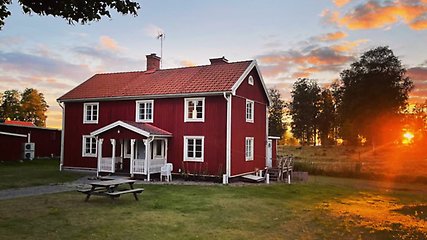 Stort rött trähus med vita knutar. Framför huset finns en liten gång och ett träbord. Bakom huset syns träd och en orange solnedgång. 