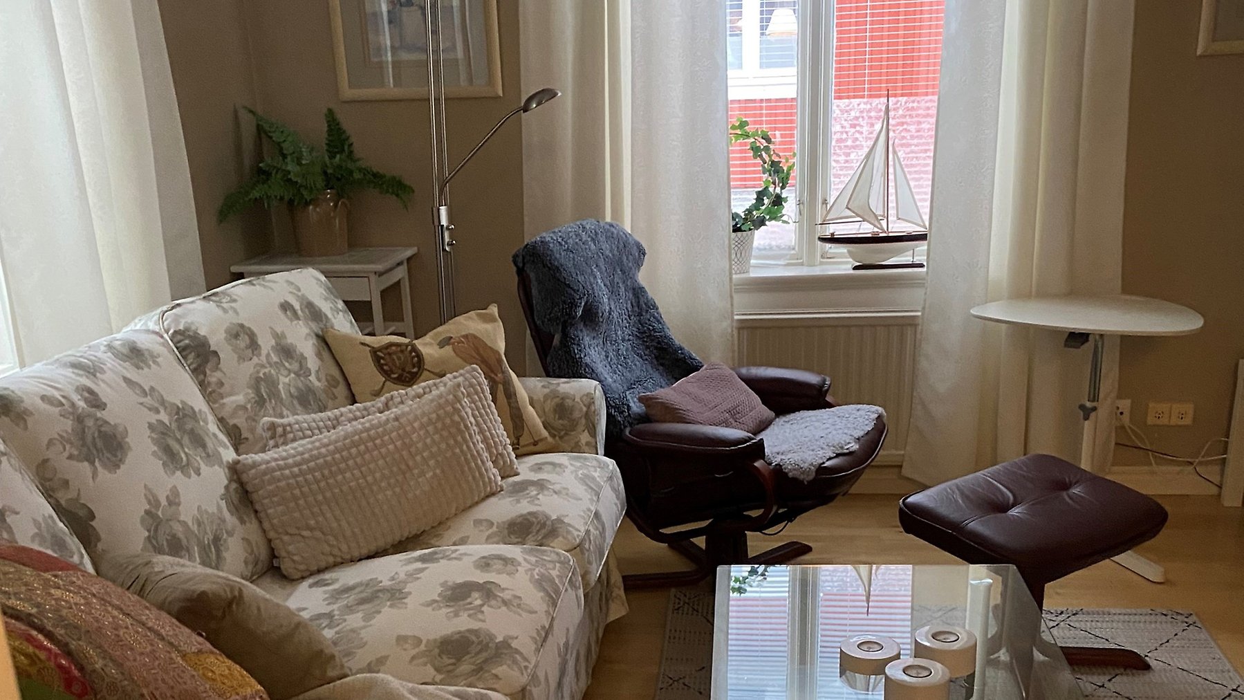 Vardagsrum med soffa, fåtölj och glasbord. Fönster med meddragna persienner i bakgrunden.