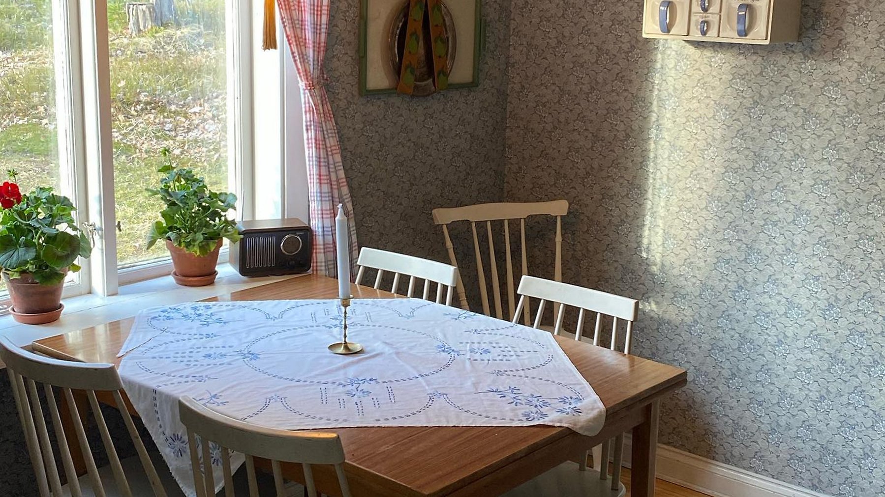 Köksbord med fyra stolar framför fönster. Borderat duk och ljusstake på bordet.
