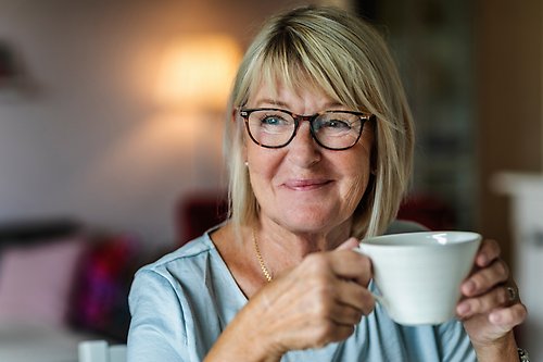 Blond kvinna med glasögon ler och håller i en kopp. Dimmig bakgrund med soffdetalj.