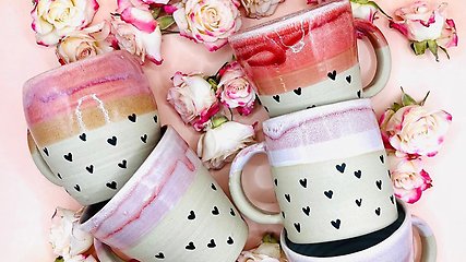 Keramik muggar i olika rosa färger och med hjärtan på. Liggandes på en rosa bakgrund med ros och vita rosor.