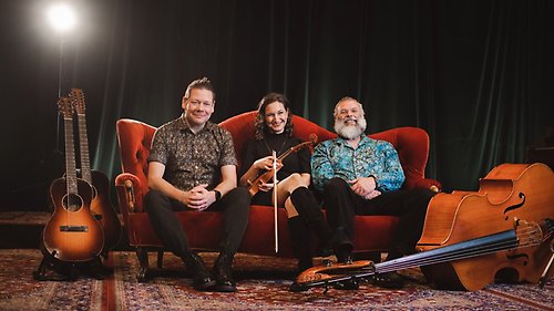 De tre musiker sitter i röd soffa med instrumenten gitarr, fiol och bas om kring sig.