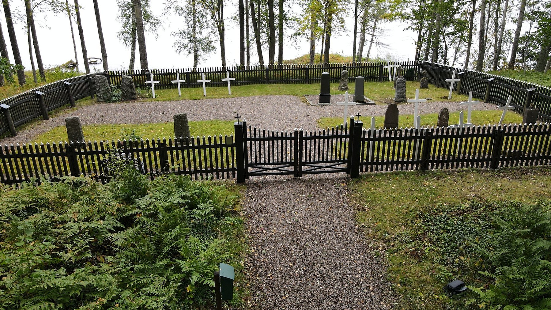Liten kyrkogård omgiven av ett svart staket. Dubbelgrind på grusgången som leder in på kyrkogården. Sjö i bakgrunden.