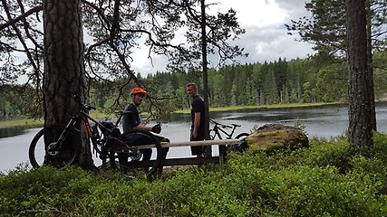 Två cyklister som tagit paus vid en bänk framför en sjö i skogen.