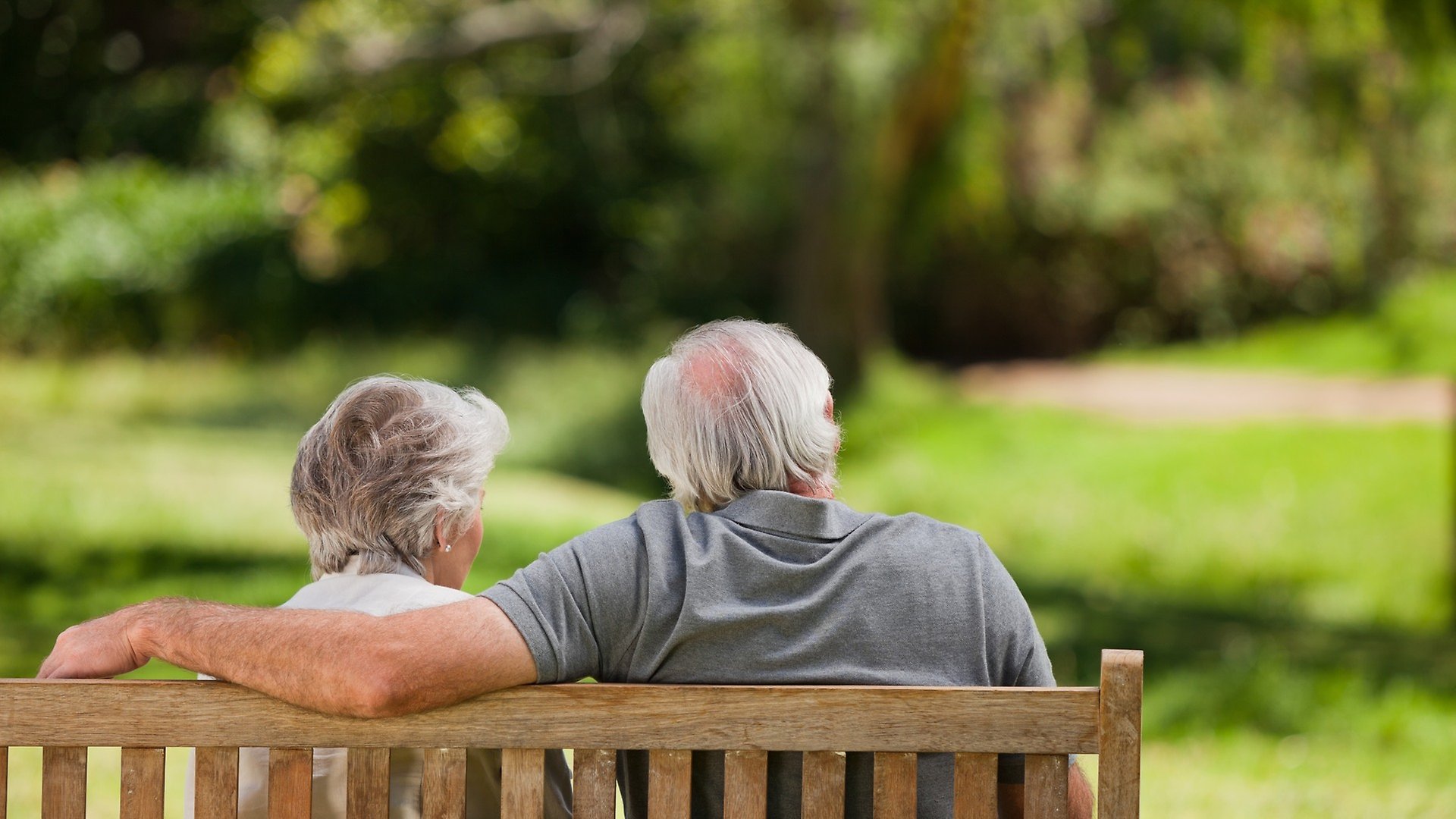Två äldre personer som sitter och håller om varandra på en bänk och blickar ut över naturen.