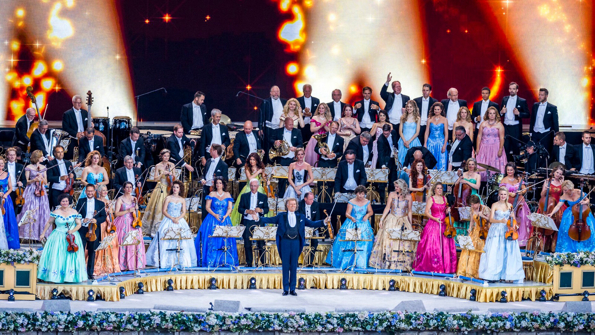 Bild över en stor scen med en orkester på. Kvinnorna i orkestern bär långa färgglada klänningar och männen bär smoking. Längst fram på scenen står dirigenten.